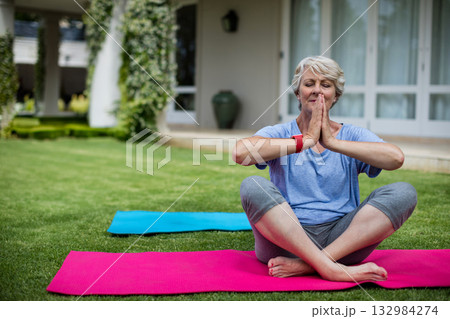 Senior woman practicing yoga on pink mat at veranda lawn with red fitness tracker, copy space Senior woman practicing yoga on pink mat at veranda lawn with red fitness tracker, copy space 132984274