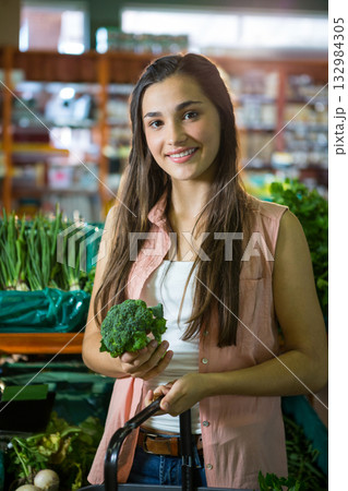 Woman holding broccoli and pushing cart in grocery produce aisle with green onions and leafy greens 132984305
