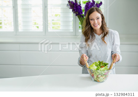 Woman holding wooden salad servers tossing veggies in glass bowl at kitchen countertop, copy space Woman holding wooden salad servers tossing veggies in glass bowl at kitchen countertop, copy space 132984311