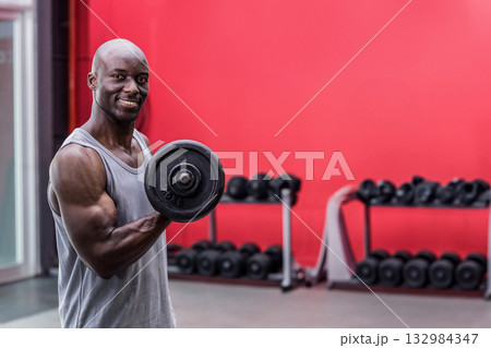 African American man performing bicep curl holding dumbbell at gym with rack, red wall, copy space 132984347