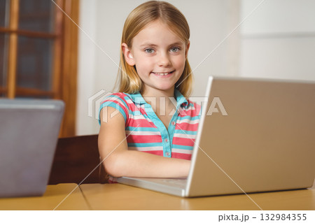 Child girl smiling directly at camera while sitting at wooden study table using silver laptop 132984355