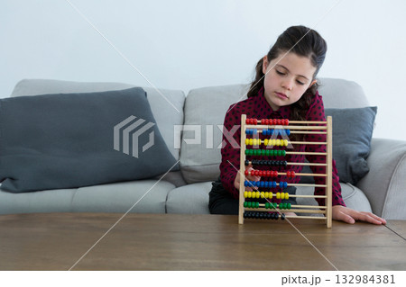 Female school-age child sitting on light grey sofa in living room using wooden abacus, copy space 132984381