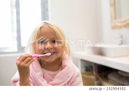 Female child brushing teeth at bathroom sink in pink bathrobe with pink toothbrush and gold mirror Female child brushing teeth at bathroom sink in pink bathrobe with pink toothbrush and gold mirror 132984382