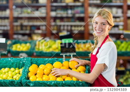 Female store employee in red apron arranging oranges in green bins at produce section, copy space Female store employee in red apron arranging oranges in green bins at produce section, copy space 132984389
