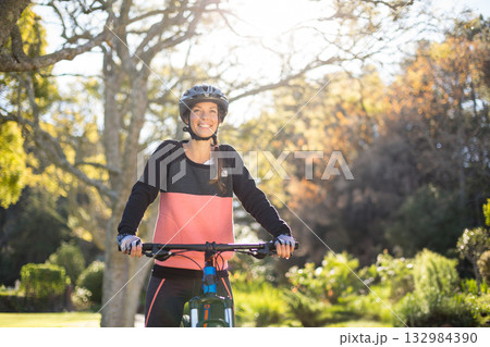 Woman wearing cycling helmet and gloves holding bicycle handlebars while riding through public park 132984390