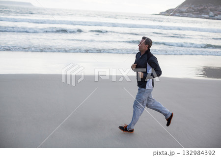 Man jogging along wet sand at shoreline wearing athletic jacket and running shoes, copy space 132984392