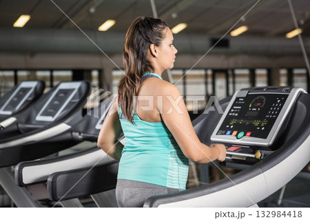 Woman in tank top and leggings standing on treadmill holding handrails checking console at gym Woman in tank top and leggings standing on treadmill holding handrails checking console at gym 132984418