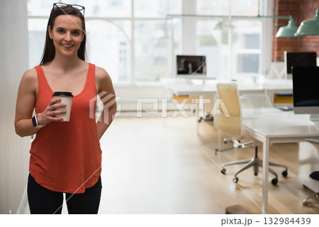Woman standing in modern office wearing orange top and smartwatch holding coffee cup, copy space 132984439