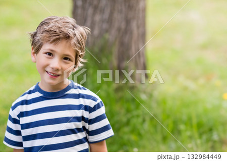 Boy standing in grassy park by tree trunk, wearing navy and white striped T-shirt and smiling 132984449