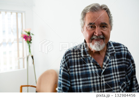Senior man standing in living room wearing checkered shirt beside vase with flowers, copy space 132984456