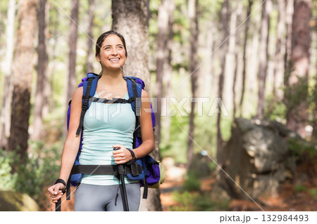 Woman standing and looking upward on sunlit dirt forest trail holding trekking poles and backpack Woman standing and looking upward on sunlit dirt forest trail holding trekking poles and backpack 132984493