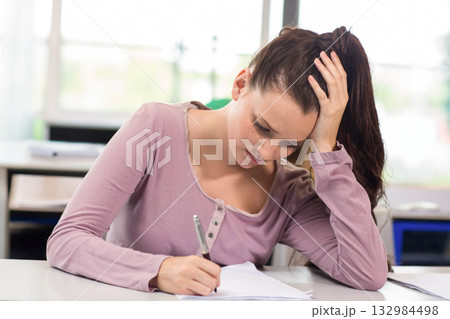 Woman wearing mauve long-sleeve top writing with pen on paper at classroom desk while resting head 132984498
