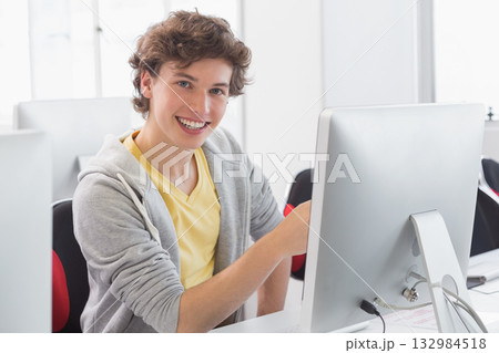 Male student sitting at white desk in computer lab operating silver monitor and typing on keyboard Male student sitting at white desk in computer lab operating silver monitor and typing on keyboard 132984518