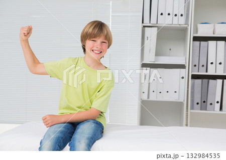 School-age boy flexing right arm while sitting on exam table under window blinds in exam room School-age boy flexing right arm while sitting on exam table under window blinds in exam room 132984535