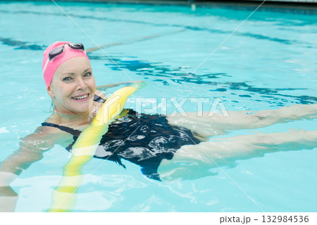 Senior woman floating on yellow noodle at outdoor pool wearing black swimsuit pink swim cap goggles 132984536