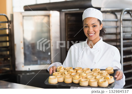 African American woman pastry chef holding tray of golden pastries at bakery prep table, copy space 132984537