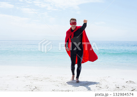 Mid adult man in black bodysuit and red cape and eye-mask standing on beach raising fist Mid adult man in black bodysuit and red cape and eye-mask standing on beach raising fist 132984574