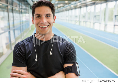 Male athlete standing arms crossed on track at sports facility wearing earphones and armband Male athlete standing arms crossed on track at sports facility wearing earphones and armband 132984575