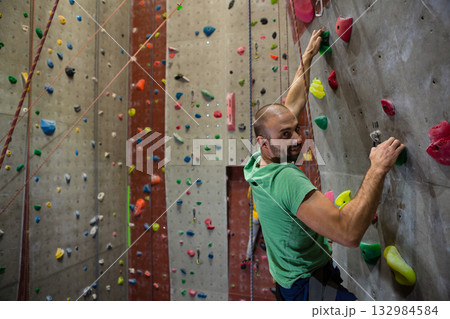 Man climbing artificial rock wall in gym gripping multicolored holds with rope harness, copy space 132984584
