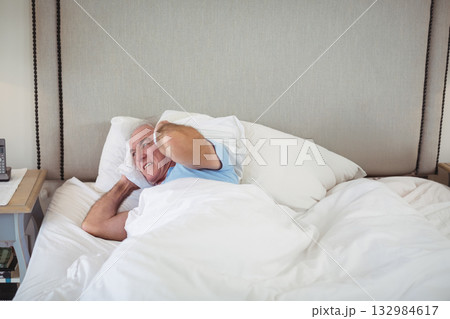 Senior man lying in bedroom pressing pillow over ears beside nightstand with telephone, copy space Senior man lying in bedroom pressing pillow over ears beside nightstand with telephone, copy space 132984617