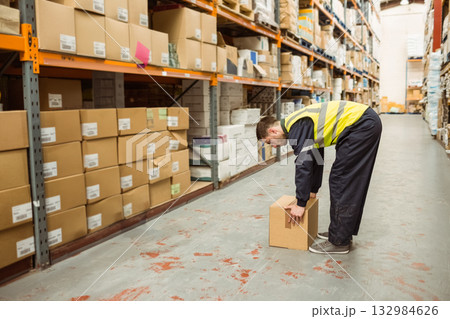Male worker in high-visibility vest bending and lifting cardboard box on aisle with shelving racks Male worker in high-visibility vest bending and lifting cardboard box on aisle with shelving racks 132984626