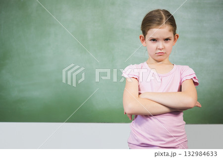 Schoolgirl standing with crossed arms in classroom at chalk tray under green chalkboard, copy space 132984633