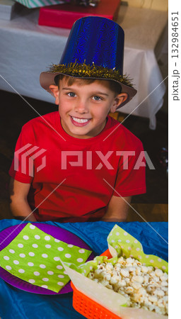 School-age boy sitting at party table wearing red shirt and blue top hat with popcorn basket 132984651