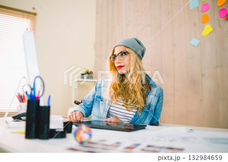 Graphic designer wearing glasses and beanie sketching color art on tablet at studio desk Graphic designer wearing glasses and beanie sketching color art on tablet at studio desk 132984659
