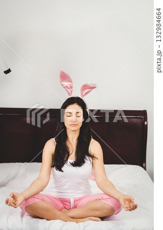Asian woman sitting cross legged meditating on bed with white sheets wearing pink bunny ears Asian woman sitting cross legged meditating on bed with white sheets wearing pink bunny ears 132984664
