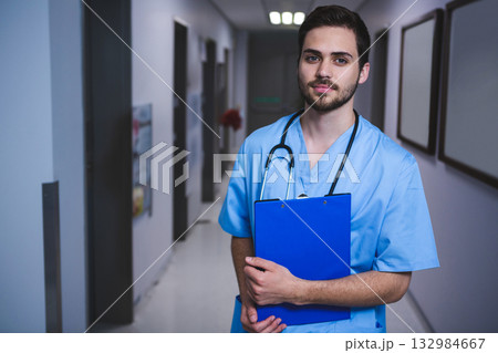 Male health worker standing in corridor holding blue clipboard and wearing stethoscope, copy space 132984667