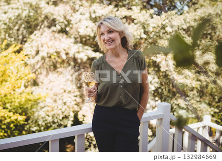 Woman standing on wooden deck in backyard holding wine glass and leaning on railing Woman standing on wooden deck in backyard holding wine glass and leaning on railing 132984669