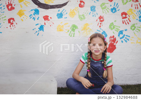 Child girl sitting cross-legged on grass in front of handprint wall holding red paint, copy space Child girl sitting cross-legged on grass in front of handprint wall holding red paint, copy space 132984688