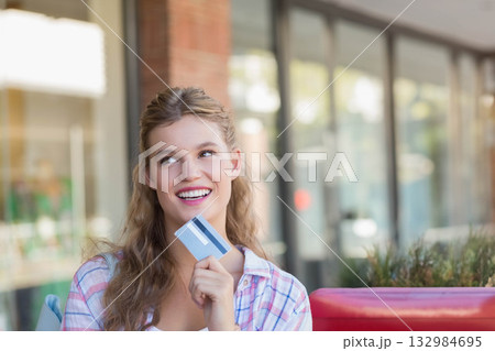 Woman sitting on red bench in front of glass storefront windows holding credit card and smiling 132984695