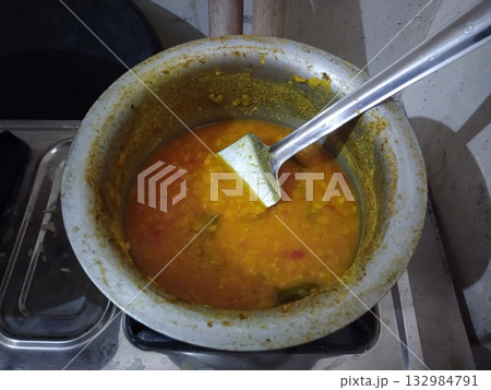 Traditional Indian kitchen scene with lentils being cooked in a steel pot on a gas stove, simple rural home meal preparation showcasing authentic daily food culture 132984791