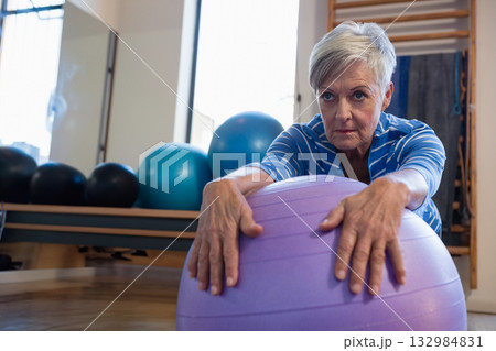 Senior woman leaning forward over purple stability ball inside fitness studio with wall bars Senior woman leaning forward over purple stability ball inside fitness studio with wall bars 132984831