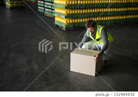 Man in safety vest crouching and lifting box by pallets of bottles in warehouse, copy space 132984838
