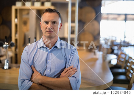 Middle-aged man standing with arms crossed inside brewery taproom by metal beer taps and barrels Middle-aged man standing with arms crossed inside brewery taproom by metal beer taps and barrels 132984846
