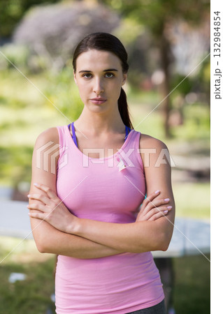 Woman standing in park wearing pink tank top with breast cancer ribbon, holding crossed arms Woman standing in park wearing pink tank top with breast cancer ribbon, holding crossed arms 132984854
