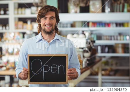Adult man standing inside cafe holding chalkboard sign while showcasing espresso machine and jars Adult man standing inside cafe holding chalkboard sign while showcasing espresso machine and jars 132984870