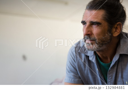 Mature man sitting in home studio gazing toward right side at blank off-white wall, copy space Mature man sitting in home studio gazing toward right side at blank off-white wall, copy space 132984881