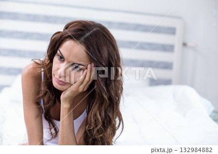 Woman sitting on bed wearing white tank top and gazing past grey striped headboard 132984882