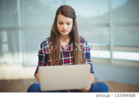 Woman sitting cross-legged on bench holding silver laptop in lounge with roller-shade windows 132984886