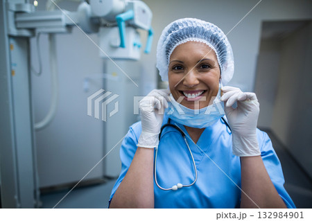 Female nurse pulling down surgical mask with gloved hands in radiology room with X-ray machine Female nurse pulling down surgical mask with gloved hands in radiology room with X-ray machine 132984901