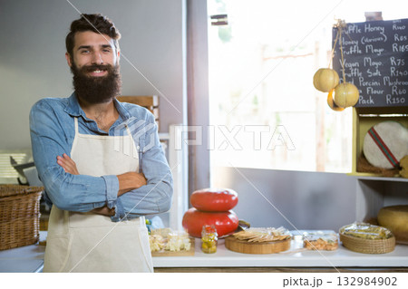Male shopkeeper in apron smiling and showing cheese wheels and olives in cheese shop, copy space 132984902