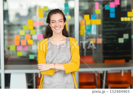 Female professional standing smiling by glass wall with sticky notes beside meeting table in office Female professional standing smiling by glass wall with sticky notes beside meeting table in office 132984903