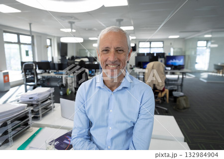 Senior man sitting on white desk in open-plan office smiling with laptop documents glass of water Senior man sitting on white desk in open-plan office smiling with laptop documents glass of water 132984904