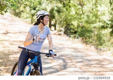 Asian woman pausing on sunlit forest trail holding sunglasses, wearing helmet and gripping bike Asian woman pausing on sunlit forest trail holding sunglasses, wearing helmet and gripping bike 132984910