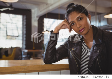 Indian woman resting head on hand at café counter wearing leather jacket, striped shirt, copy space 132984915