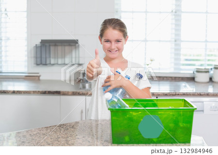 Girl standing at countertop holding empty bottles near green recycle bin giving thumbs up at home 132984946