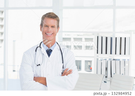Male doctor standing in exam room wearing lab coat with stethoscope beside binder-filled shelves 132984952